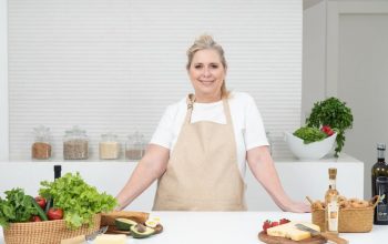 A mature woman in an apron stands at a kitchen counter with fresh produce and cheese, ready for cooking.